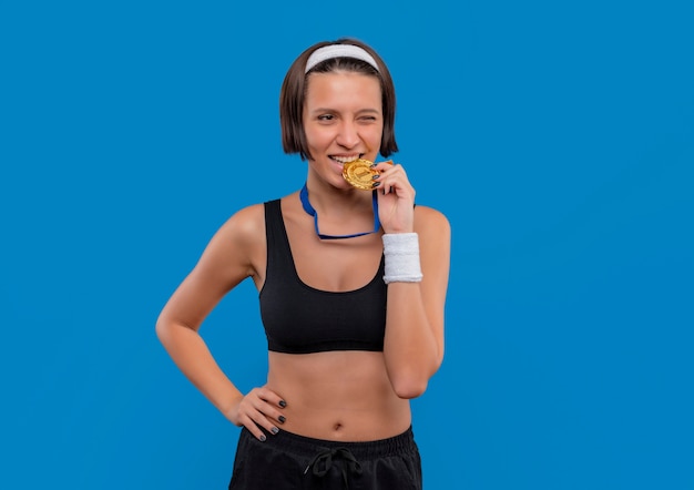 Jonge fitness vrouw in sportkleding met gouden medaille om haar nek bijten medaille glimlachend staande over blauwe muur