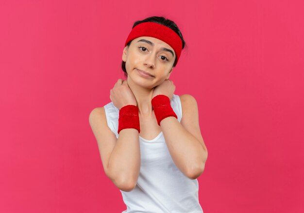 Jonge fitness vrouw in sportkleding met hoofdband aanraken van haar nek ontevreden gevoel ongemak staande over roze muur