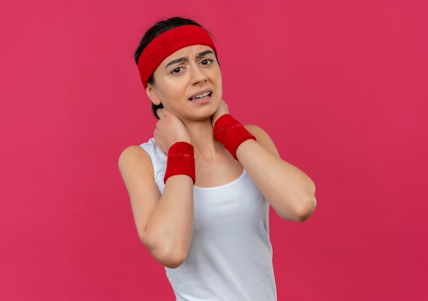Jonge fitness vrouw in sportkleding met hoofdband aanraken van haar nek ontevreden gevoel pijn staande over roze muur