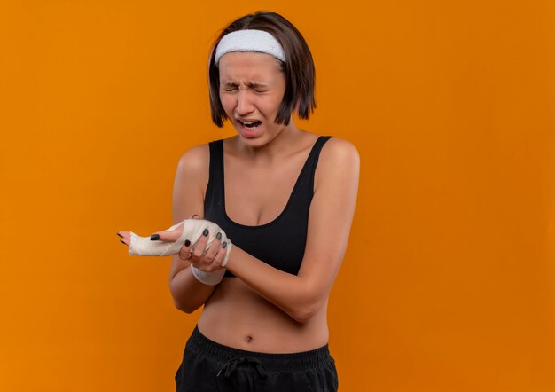 Jonge fitness vrouw in sportkleding met hoofdband aanraken van haar pols op zoek onwel lijdend aan pijn staande over oranje muur