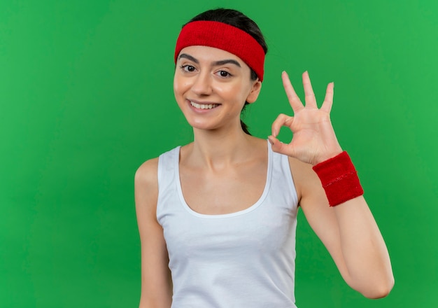 Jonge fitness vrouw in sportkleding met hoofdband gelukkig en positief glimlachend vrolijk doet ok teken staande over groene muur