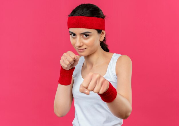 Jonge fitness vrouw in sportkleding met hoofdband glimlachend poseren als een bokser met vuisten permanent over roze muur