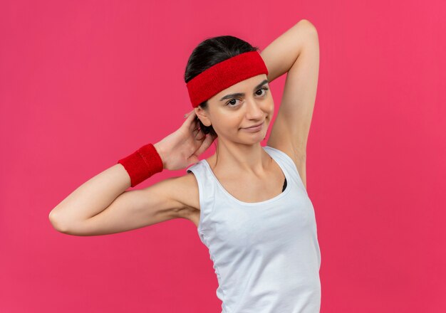 Jonge fitness vrouw in sportkleding met hoofdband glimlachend uitrekken zich staande over roze muur