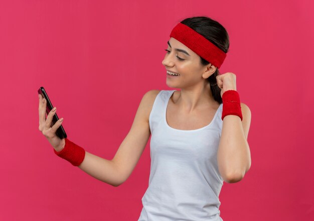 Jonge fitness vrouw in sportkleding met hoofdband houden op het scherm van haar smartphone glimlachend gebalde vuist staande over roze muur