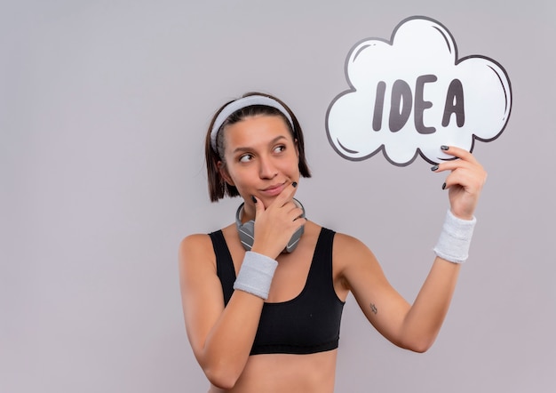 Jonge fitness vrouw in sportkleding met hoofdband houden toespraak bubble bord met woord idee opzij kijken met peinzende uitdrukking staande over witte muur