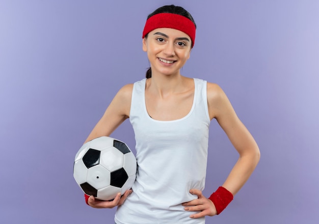 Jonge fitness vrouw in sportkleding met hoofdband houden voetbal glimlachend zelfverzekerd blij en positief staande over paarse muur