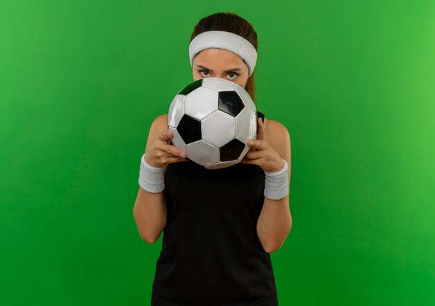 Gratis foto jonge fitness vrouw in sportkleding met hoofdband houden voetbal verbergt gezicht erachter gluren over staande over groene muur