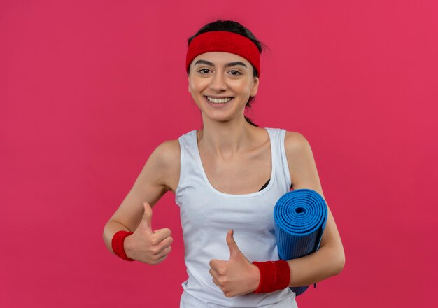 Jonge fitness vrouw in sportkleding met hoofdband houden yoga mat duimen opdagen glimlachend vrolijk staande over roze muur