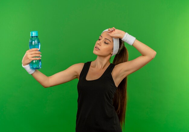 Jonge fitness vrouw in sportkleding met hoofdband met fles water op zoek moe staande over groene muur