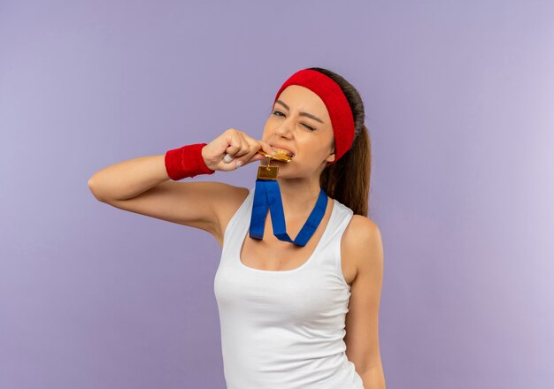Jonge fitness vrouw in sportkleding met hoofdband met gouden medaille om haar nek haar medaille bijten op zoek zelfverzekerd staande over grijze muur