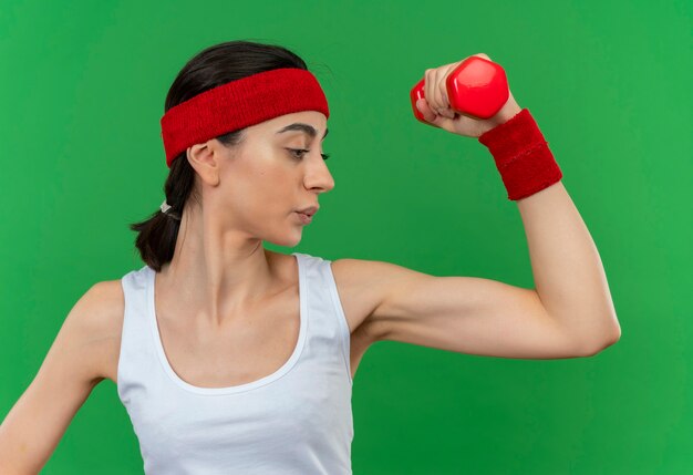 Jonge fitness vrouw in sportkleding met hoofdband met halter hand opheffen doen oefeningen gespannen opzij kijken staande over groene muur