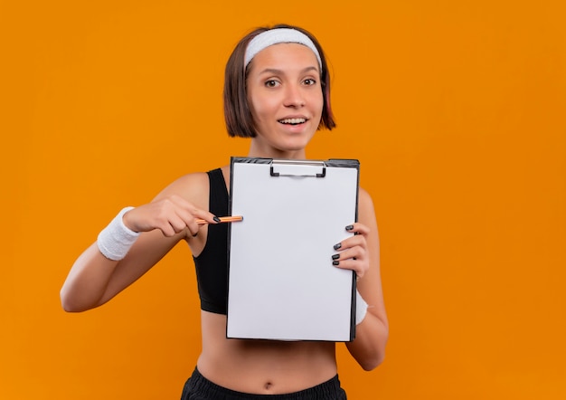 Jonge fitness vrouw in sportkleding met hoofdband met klembord met blanco pagina's wijzend met pen naar het glimlachend vriendelijk staande over oranje muur