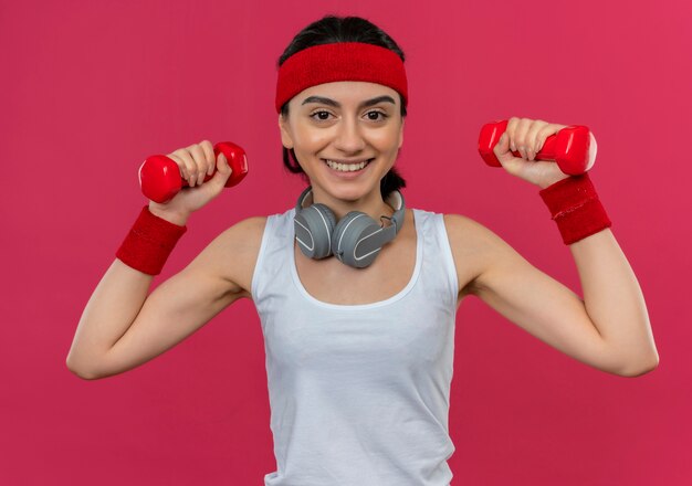 Jonge fitness vrouw in sportkleding met hoofdband met twee halters doen oefeningen met glimlach op gezicht staande over roze muur