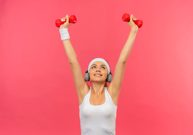 Jonge fitness vrouw in sportkleding met hoofdband met twee halters doen oefeningen op zoek zelfverzekerd staande over roze muur