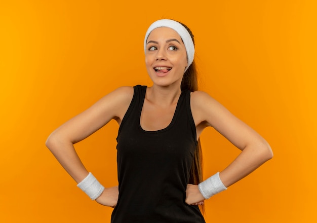 Jonge fitness vrouw in sportkleding met hoofdband opzij kijken gelukkig en positief glimlachen staande over oranje muur