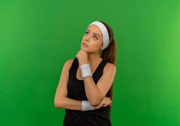 Jonge fitness vrouw in sportkleding met hoofdband opzij kijken met hand op kin met peinzende uitdrukking staande over groene muur