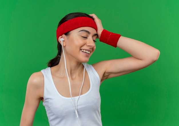 Jonge fitness vrouw in sportkleding met hoofdband opzij kijken verward glimlachend staande over groene muur