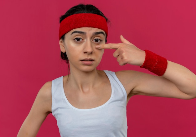 Gratis foto jonge fitness vrouw in sportkleding met hoofdband verward wijzend met wijsvinger naar haar neus staande over roze muur