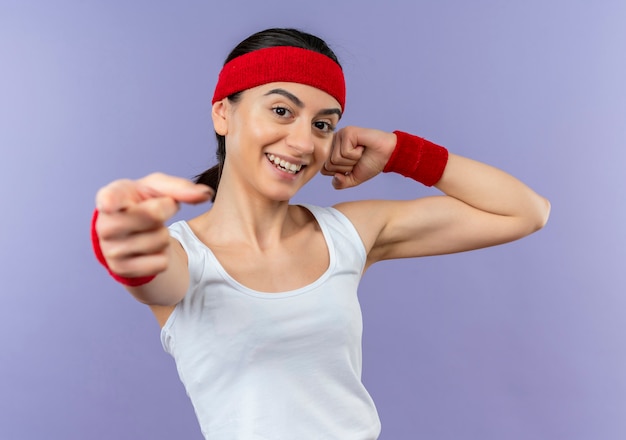 Jonge fitness vrouw in sportkleding met hoofdband vuist opheffen wijzend met vinger naar camera glimlachend zelfverzekerd staande over paarse muur