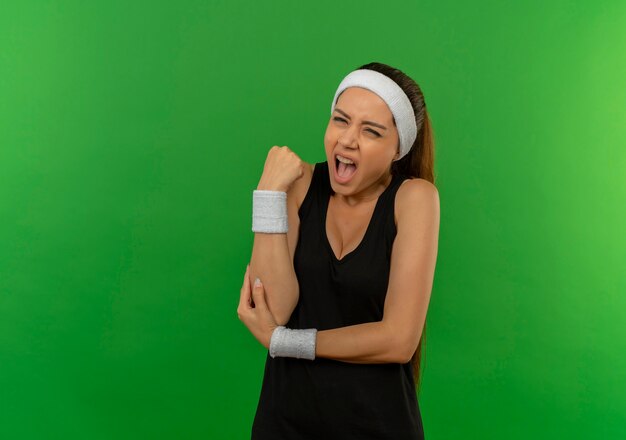Jonge fitness vrouw in sportkleding met hoofdband wat betreft elleboog die pijn heeft die zich over groene muur bevindt
