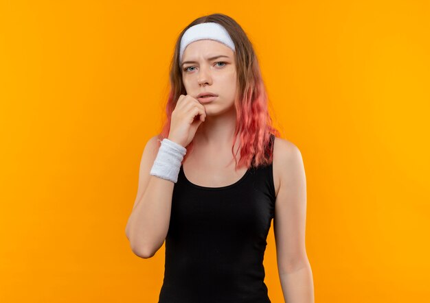 Jonge fitness vrouw in sportkleding met peinzende uitdrukking op gezicht, staande denken over oranje muur