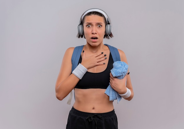 Jonge fitness vrouw in sportkleding met rugzak en koptelefoon op hoofd met handdoek met hand op haar borst bang staande over witte muur
