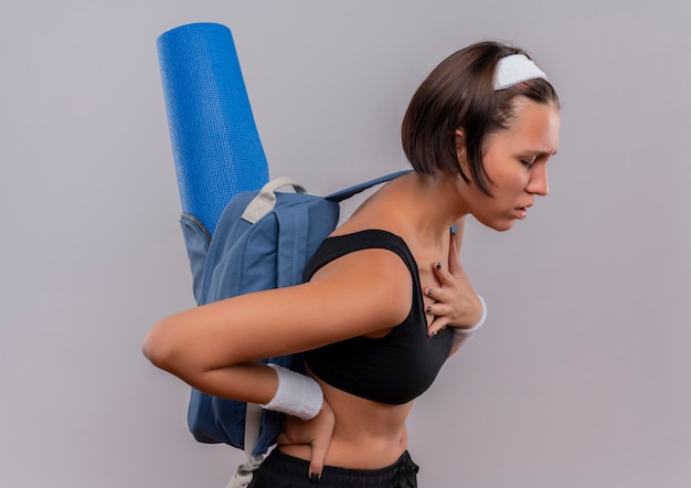 Jonge fitness vrouw in sportkleding met rugzak en yoga mat op zoek onwel hand op haar borst moe staande over witte muur