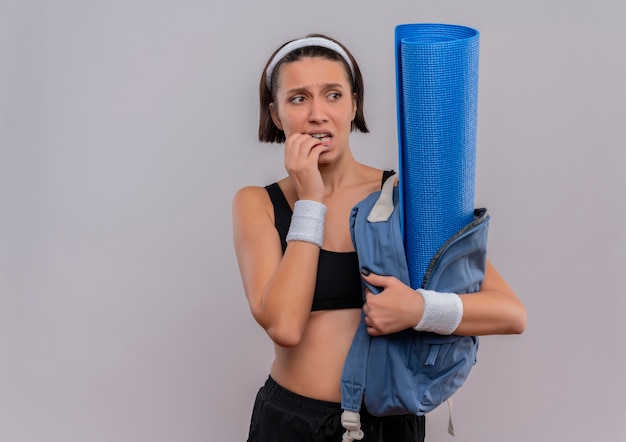 Jonge fitness vrouw in sportkleding met rugzak met yogamat opzij kijken beklemtoonde en nerveuze bijtende nagels staande over witte muur