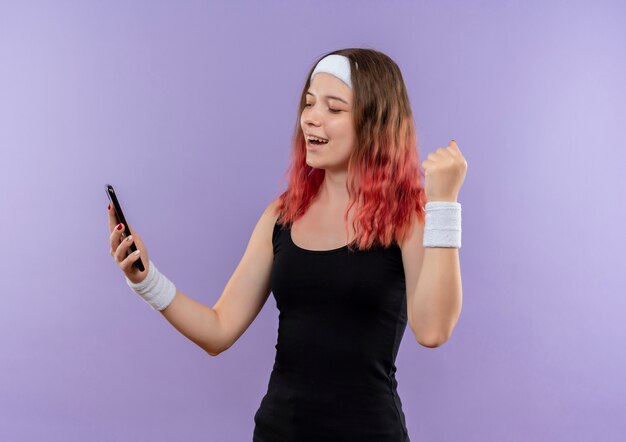 Jonge fitness vrouw in sportkleding met smartphone gebalde vuist blij en verlaten staande over paarse muur