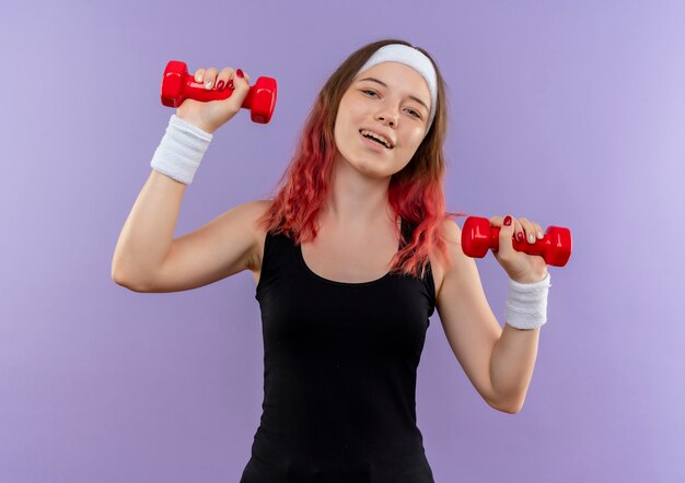 Jonge fitness vrouw in sportkleding oefeningen doen met dumbbellss glimlachend vrolijk staande over paarse muur