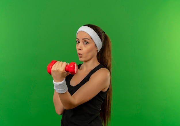 Jonge fitnessvrouw in sportkleding met hoofdband die twee domoren houdt die oefeningen doen die verward en verbaasd over groene muur kijken