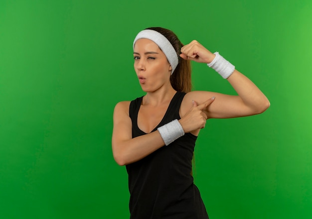 Gratis foto jonge fitnessvrouw in sportkleding met hoofdband die vuist opheft die haar biceps toont die zelfverzekerd kijken knipogend status over groene muur