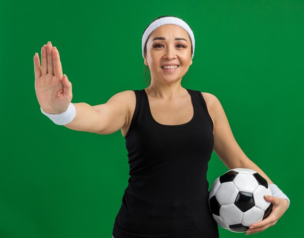 Jonge fitnessvrouw met hoofdband die voetbal vasthoudt met een glimlach op het gezicht en een stopgebaar maakt met open hand die over de groene muur staat