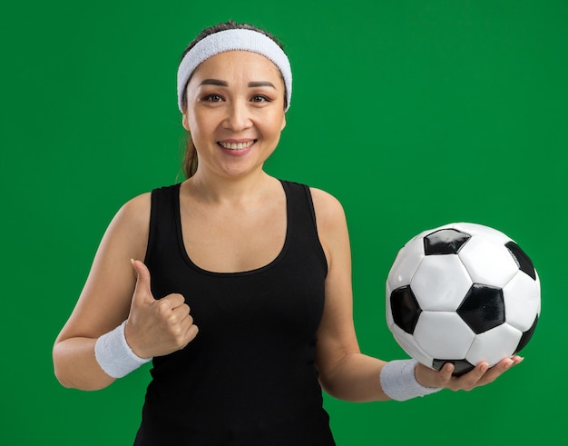 Jonge fitnessvrouw met hoofdband die voetbal vasthoudt met een glimlach op het gezicht over een groene muur