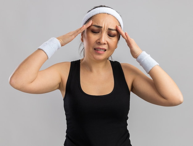 Jonge fitnessvrouw met hoofdband en armbanden die haar hoofd aanraakt en er onwel uitziet met hoofdpijn die over een witte muur staat