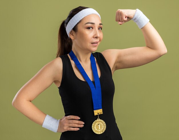 Jonge fitnessvrouw met hoofdband en armbanden met gouden medaille om haar nek die er zelfverzekerd uitziet met een vuist die biceps toont