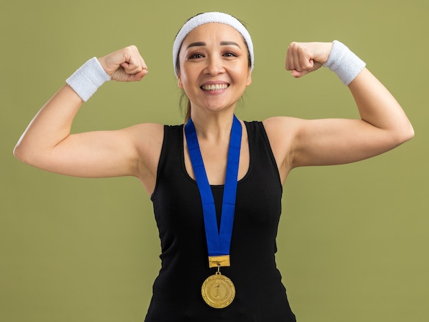 Gratis foto jonge fitnessvrouw met hoofdband en armbanden met gouden medaille om haar nek glimlachend zelfverzekerde vuisten die kracht en macht tonen die over groene muur staan