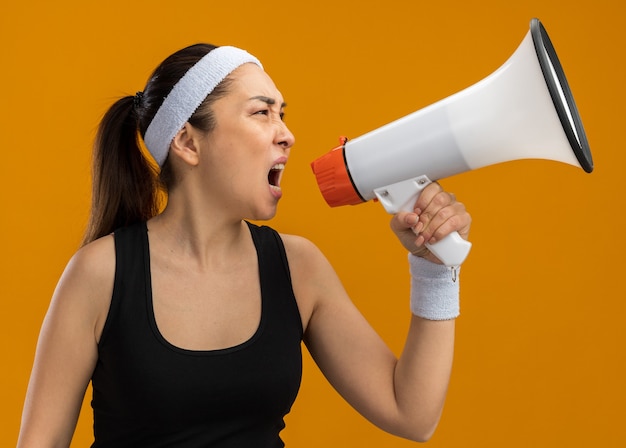 Jonge fitnessvrouw met hoofdband en armbanden schreeuwend naar megafoon met agressieve uitdrukking