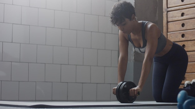 Jonge fitte en versterkte vrouw van gemengd ras die tijdens zonnige dag fitnesstraining doet met buikwiel op zolder