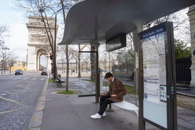 Jonge franse man wacht op het station op de bus en gebruikt zijn smartphone