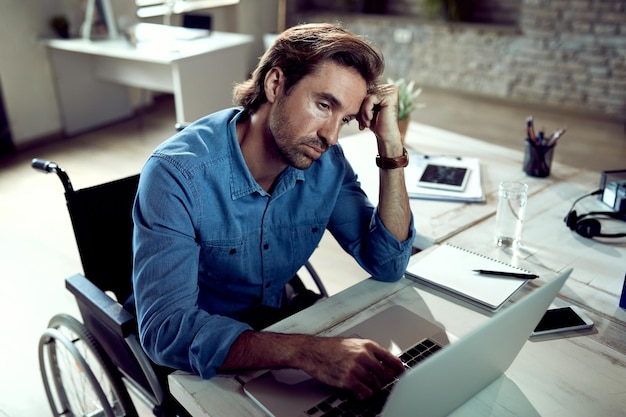 Jonge freelance werknemer in rolstoel die aan iets denkt terwijl hij op een laptop op kantoor werkt