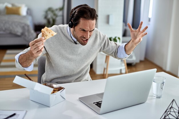 Jonge freelance werknemer voelt zich overstuur tijdens het eten en lezen van slecht nieuws via laptop thuis