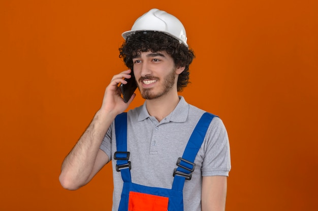 Jonge gebaarde knappe bouwer die eenvormige bouw dragen en veiligheidshelm die op mobiele telefoon spreken die met gelukkig gezicht over geïsoleerde oranje muur glimlachen