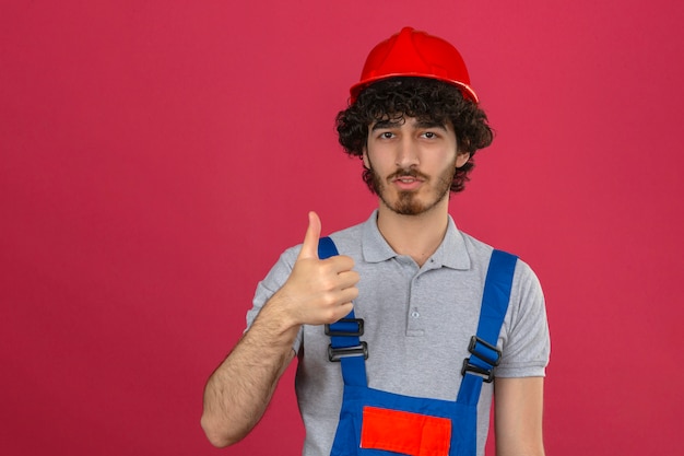 Gratis foto jonge gebaarde knappe bouwer die eenvormige bouw dragen en veiligheidshelm die tonend duim over geïsoleerde roze muur glimlachen