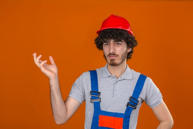 Jonge gebaarde knappe bouwer die eenvormige bouw dragen en veiligheidshelm die verward gebaar met opgeheven handuitdrukking maken als vraag over geïsoleerde oranje muur