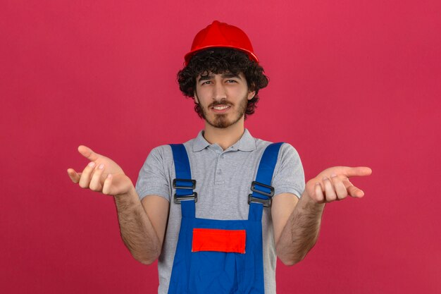 Gratis foto jonge gebaarde knappe bouwer die eenvormige bouw dragen en veiligheidshelm die zich met verwarde uitdrukking met armen en handen bevinden die over geïsoleerde roze muur worden opgeheven