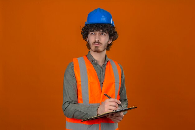 Jonge gebaarde knappe ingenieur die bouwvest en het klembord dragen die van de veiligheidshelm iets schrijven die opzij nadenkende uitdrukking op gezicht over geïsoleerde oranje muur kijken