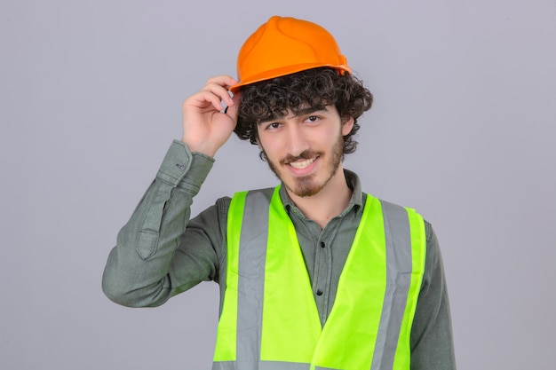 Jonge gebaarde knappe ingenieur die groetgebaar maken wat betreft zijn helm glimlachen die zich over geïsoleerde witte muur bevinden