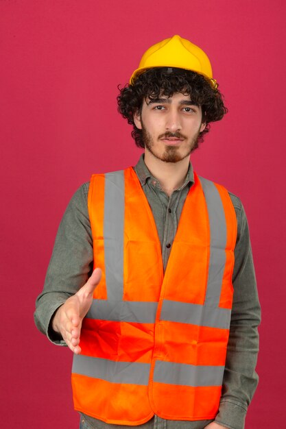 Jonge gebaarde knappe ingenieur veiligheidshelm dragen en vest die groetgebaar maken die hand aanbieden die camera met glimlach over geïsoleerde roze muur aanbieden