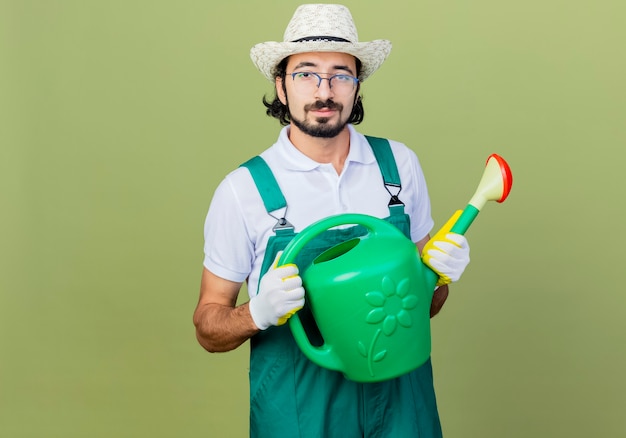 Jonge, gebaarde tuinmanmens die jumpsuit en hoed draagt die gieter houdt die voorzijde glimlachend zelfverzekerd over lichtgroene muur kijkt